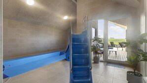 Un espace piscine avec un toboggan bleu, un patio couvert avec des meubles d’extérieur et une vue sur la campagne.