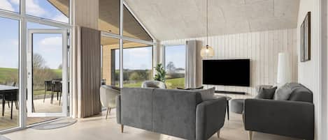 Uma sala de estar ampla com uma TV grande, sofás cinzas e vista para o exterior.