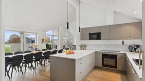Een moderne keuken met grijze kastjes, een zwarte magnetron en een inbouwoven. Er is een eethoek met zwarte stoelen en een tafel, en een groot raam met uitzicht op de omgeving.