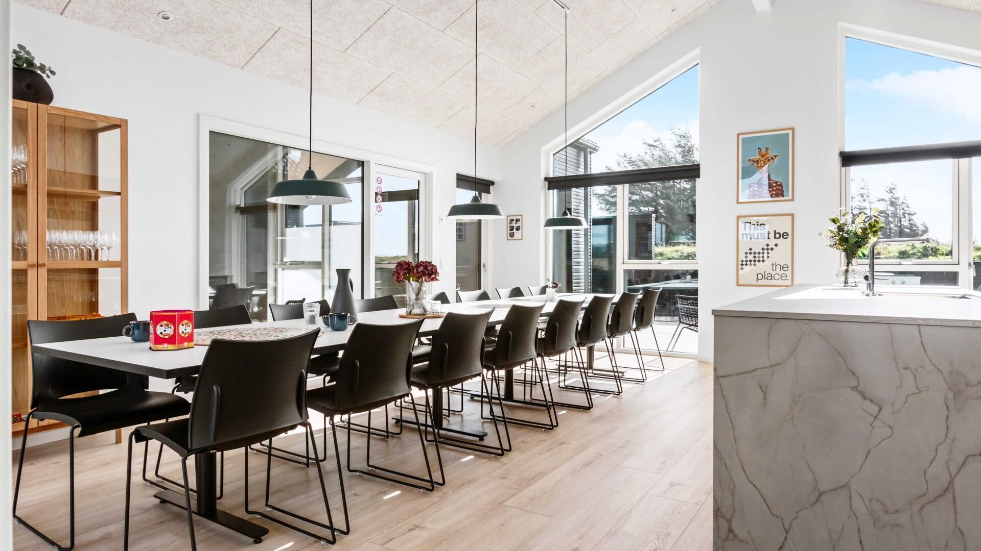 Un comedor moderno con una mesa larga, sillas negras y lámparas colgantes. Se aprecia un aparador de madera con cristalería a la izquierda y un ventanal que proporciona luz natural.