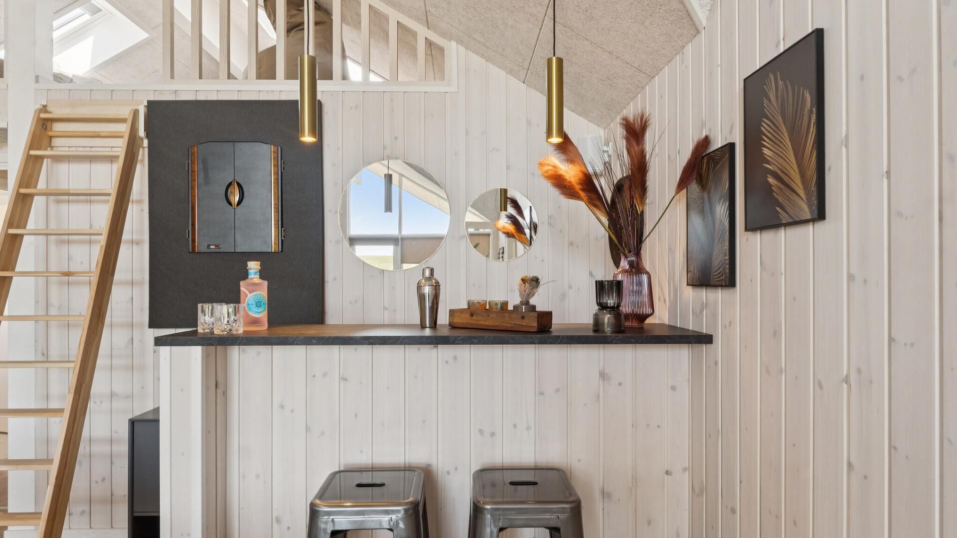 Een moderne keuken met houten paneelwanden, een zwart aanrecht, hangende pendelverlichting, een ladder, een kast, een spiegel, een vaas met gedroogde planten en twee krukken.