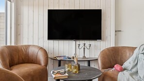 Ein Zimmer mit zwei Ledersesseln, einem runden Tisch mit Büchern, Gläsern und Dekorationsobjekten sowie einem flachbildfernseher, der an einer weißen Holzwand befestigt ist.