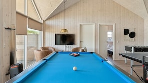 Ein Billardtisch in einem Raum mit Holzvertäfelung, darüber ein Fernseher und rechts ein Tischtennistisch.