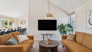 Ein modernes Wohnzimmer mit zwei Sofas, einem runden Couchtisch, einem an der Wand montierten Fernseher und einem Essbereich, der durch eine offene Tür sichtbar ist.
