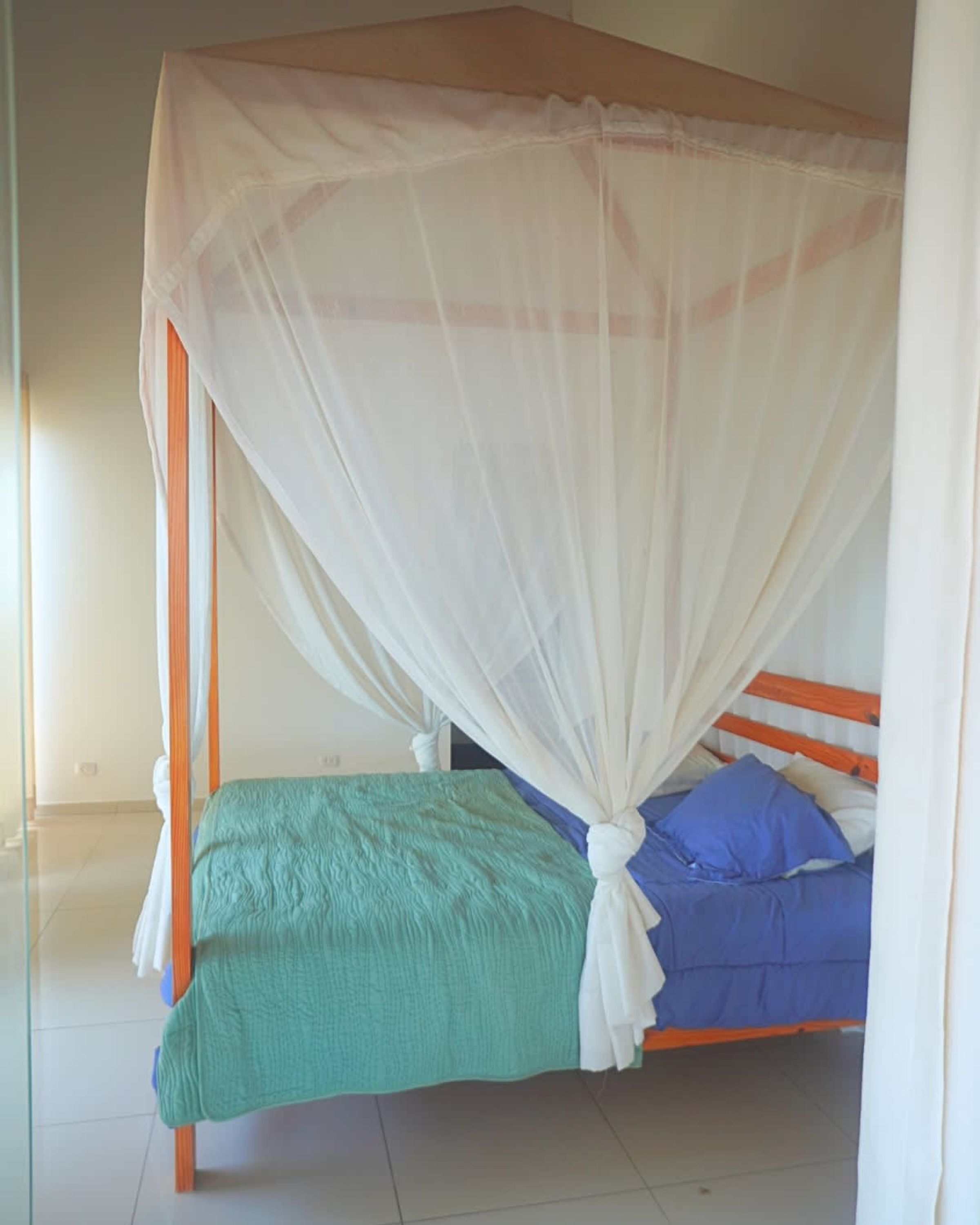 A single bed with a wooden frame, a green bedspread, and a blue pillow. A white canopy covers the bed.