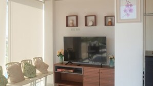 A living room with a flat-screen TV on a wooden stand, a sofa, and a coffee table. There are three framed pictures on the wall and a vase with flowers on the coffee table. - HOP Aloha Beachfront House With Direct Access (Chincha Baja)