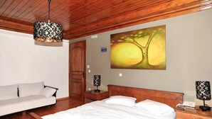 Une chambre avec un plafond en bois, un lit, une table de chevet, une lampe, un tableau représentant un arbre et un canapé.