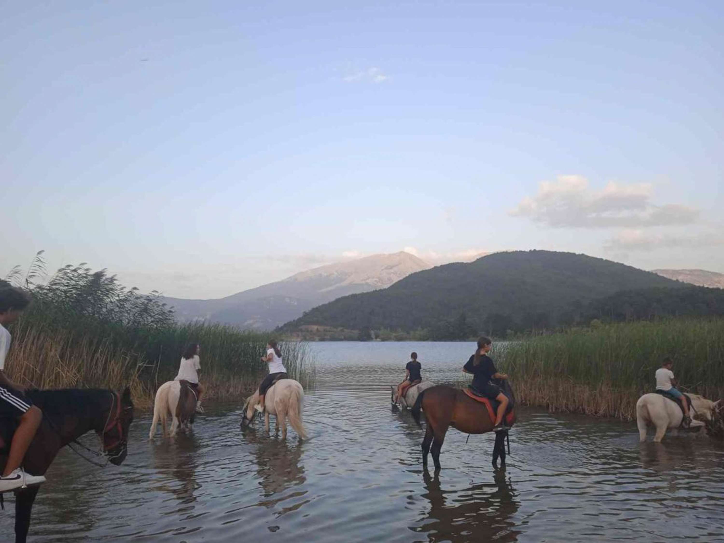 Um grupo de pessoas cavalga por águas rasas, com uma paisagem montanhosa ao fundo.