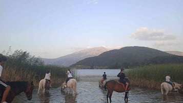 Um grupo de pessoas cavalga por águas rasas, com uma paisagem montanhosa ao fundo.