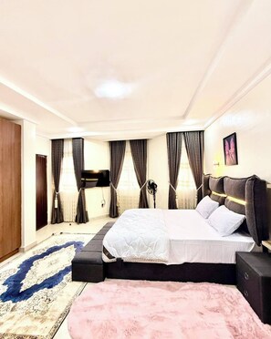 A modern bedroom with a large bed, dark curtains, a wall-mounted TV, and a decorative rug. - Declans Apartment (Abuja)