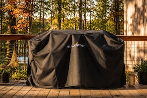 A covered grill with the Blackstone logo, set on a wooden deck with potted plants and a wooden railing.