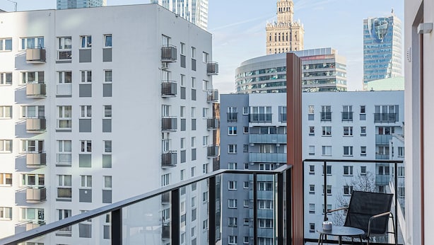 A balcony with a view of modern buildings, including a clock tower, and a chair. - RentPlanet - przy Dworcu Centralnym (Warsaw)