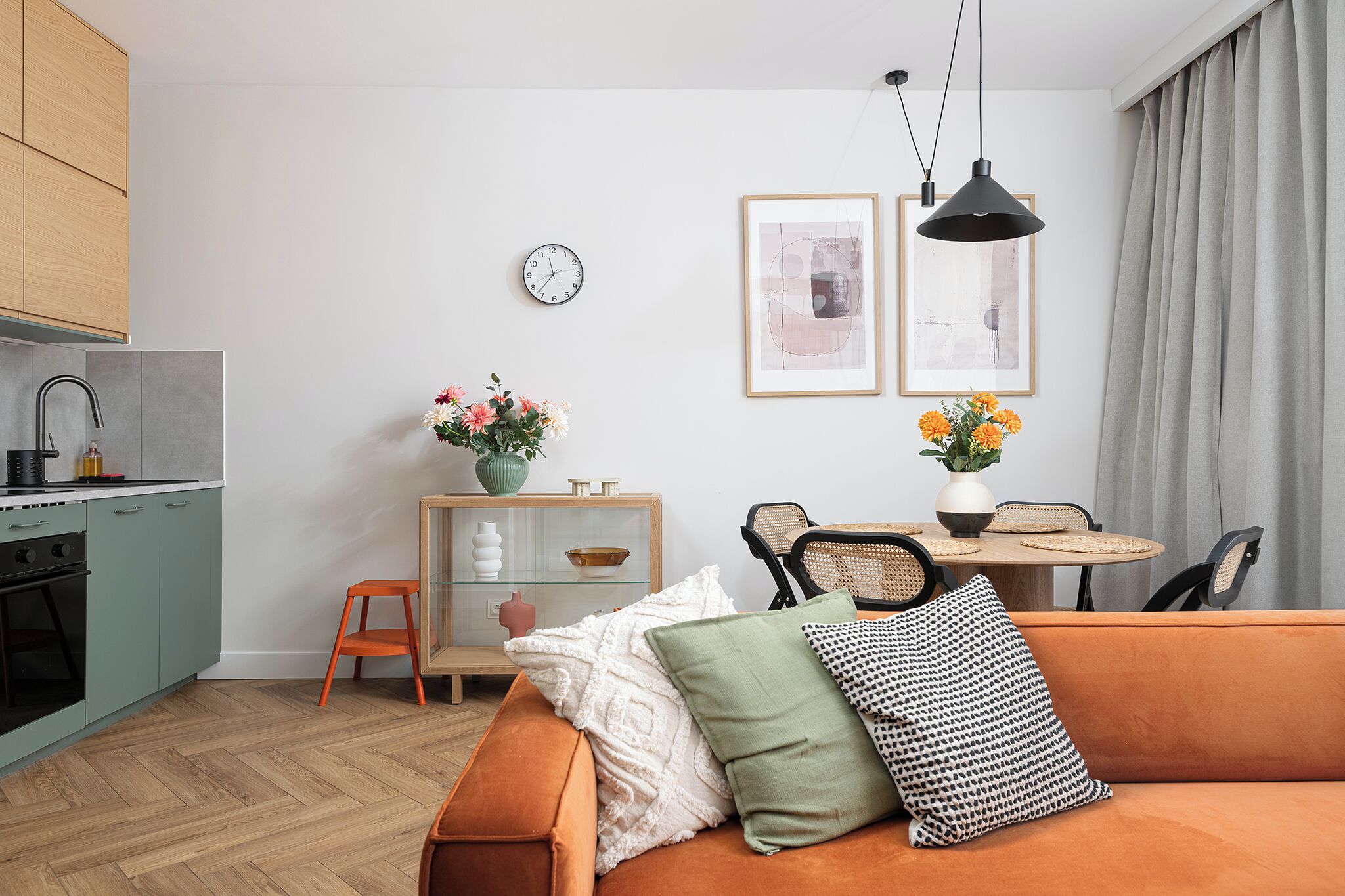 A modern living room with a sofa, a dining table with chairs, a kitchen area, and a clock on the wall.