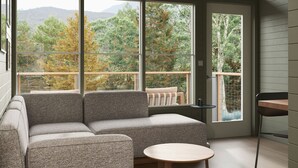 A modern living room with a sectional sofa, a round wooden table, and large windows offering a view of trees and hills. - Outbound Yosemite (Oakhurst)