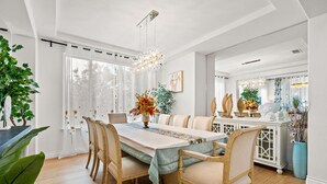Un espace repas avec une longue table, de nombreuses chaises et un lustre. On y trouve également des plantes en pot et un meuble blanc orné d’objets décoratifs.