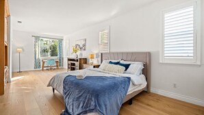 Une chambre avec un grand lit, un bureau et une chaise. Il y a une fenêtre avec des stores et une vue sur la verdure à l’extérieur.