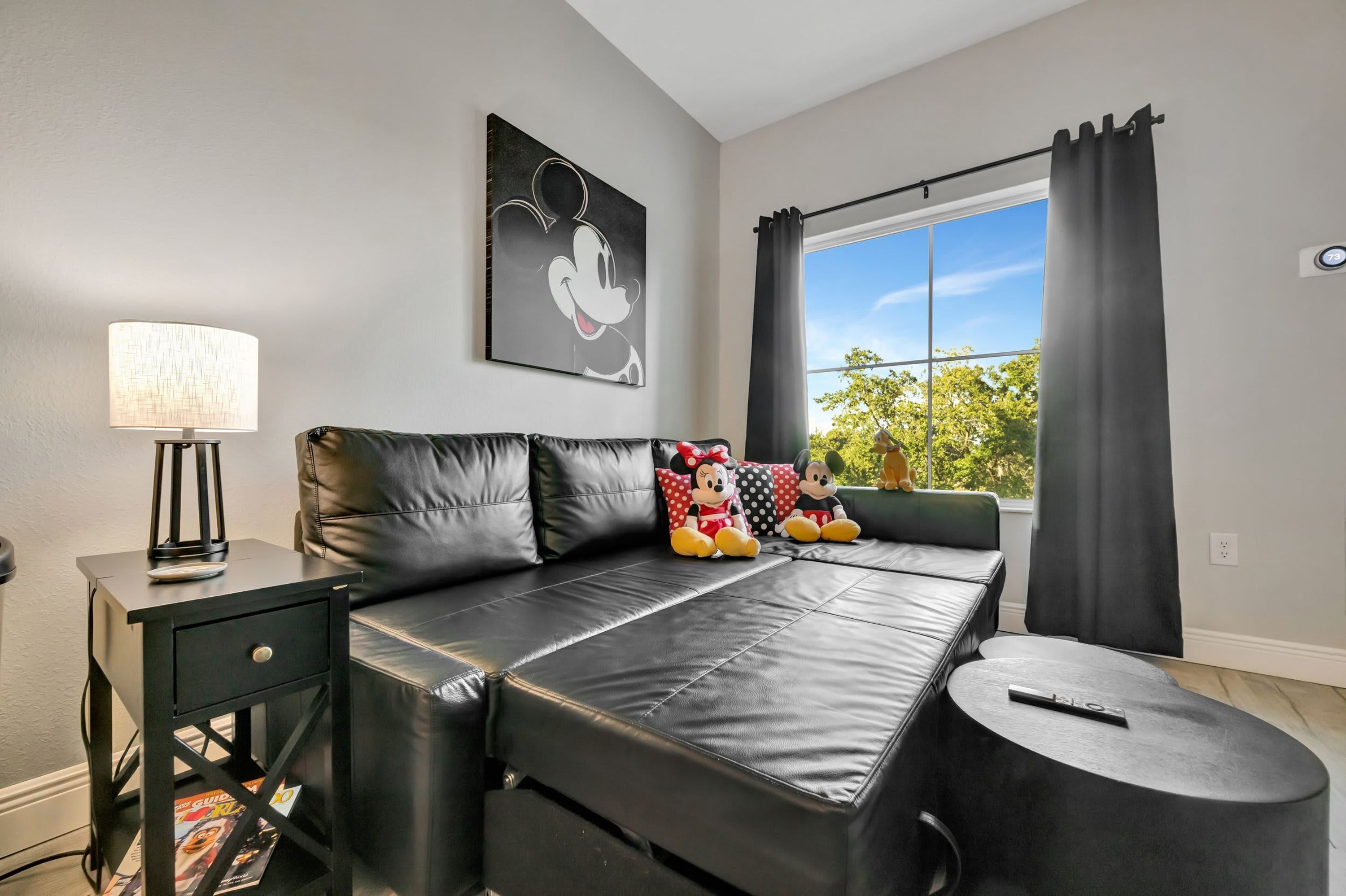 A modern living room with a black leather sofa, a side table with a lamp, and a window with curtains.