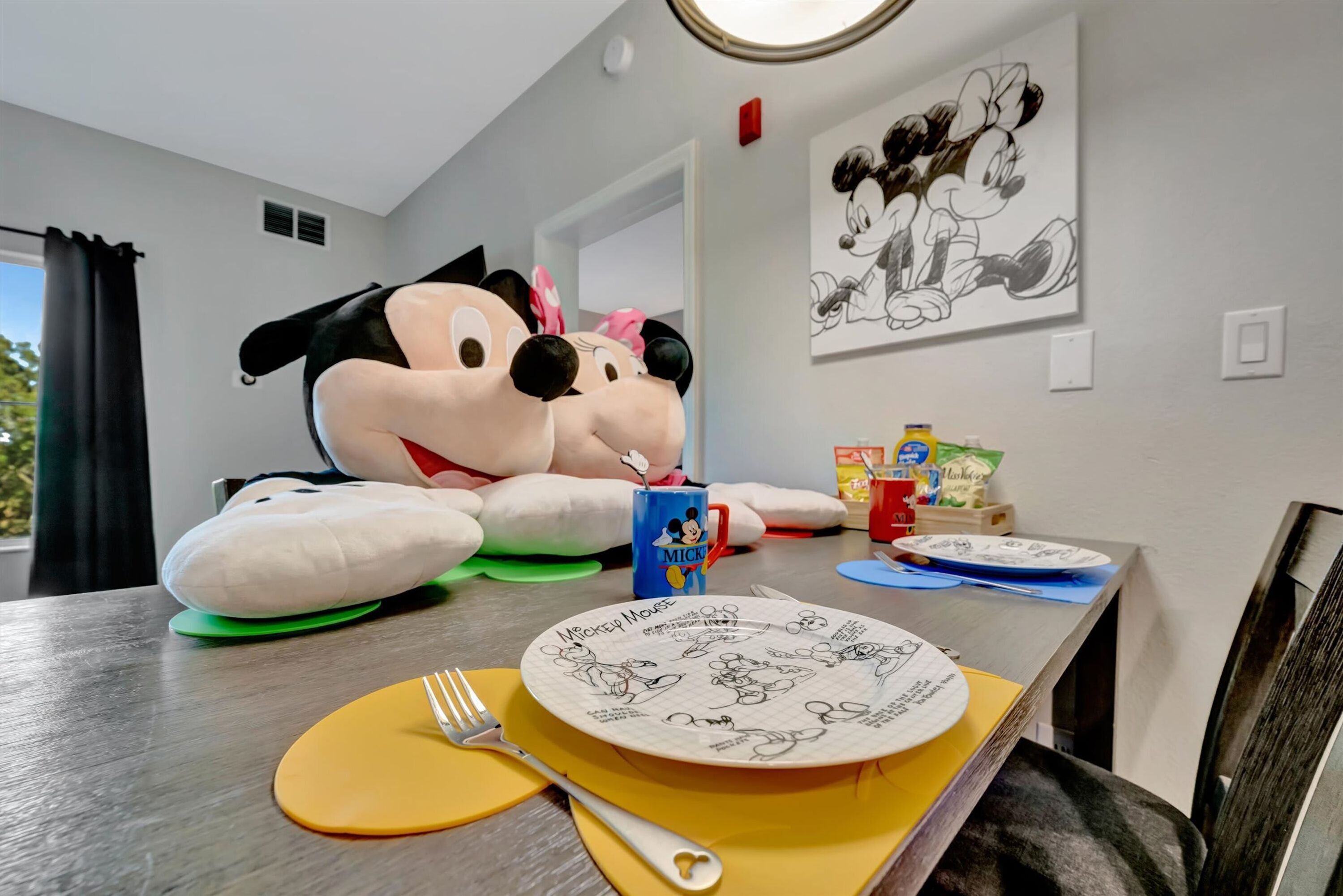 A dining table with Mickey Mouse plush toys, a Mickey Mouse mug, and plates with Mickey Mouse drawings.