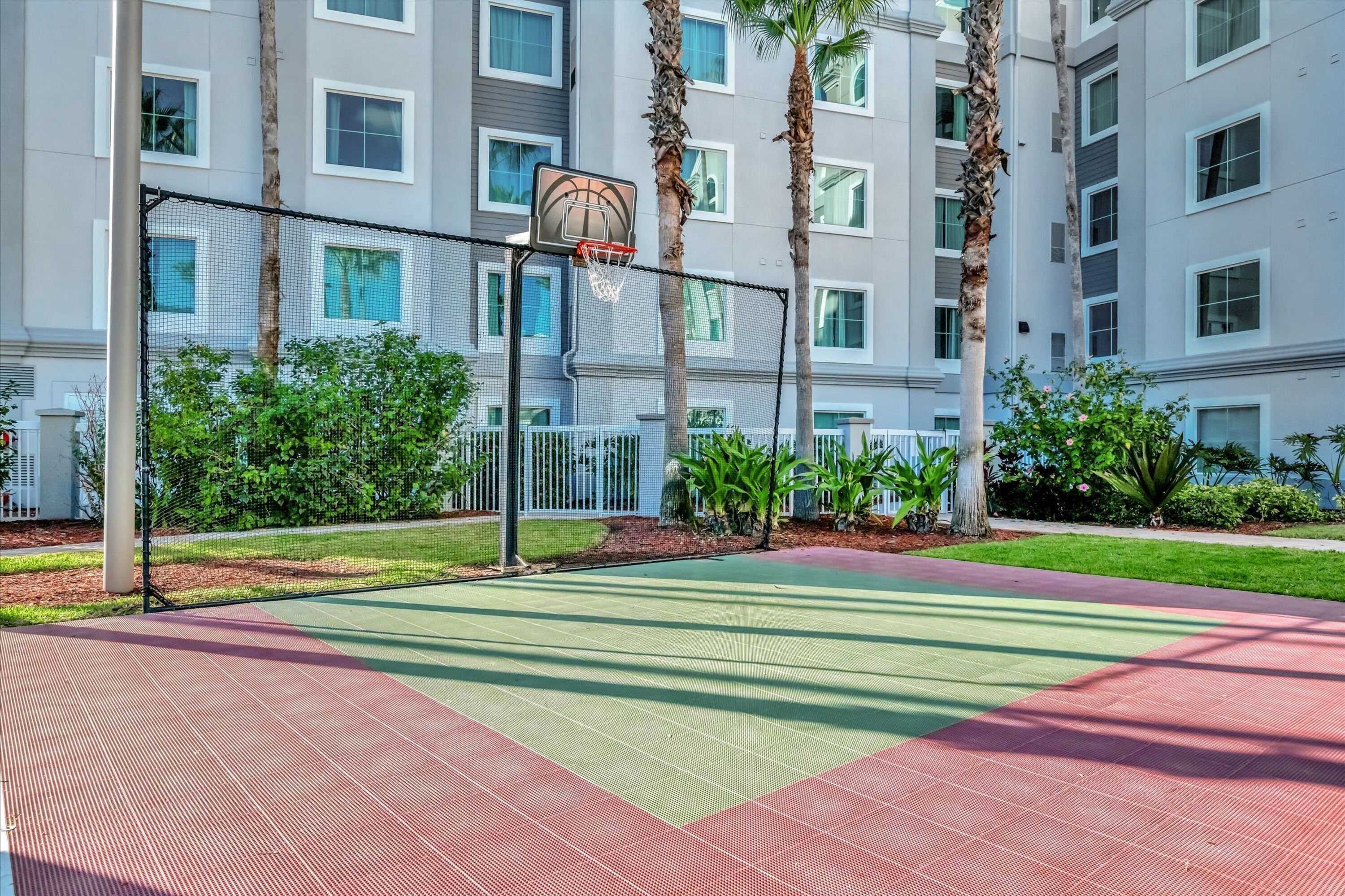 A basketball court with a net, surrounded by a fence, palm trees, and a building with multiple windows.
