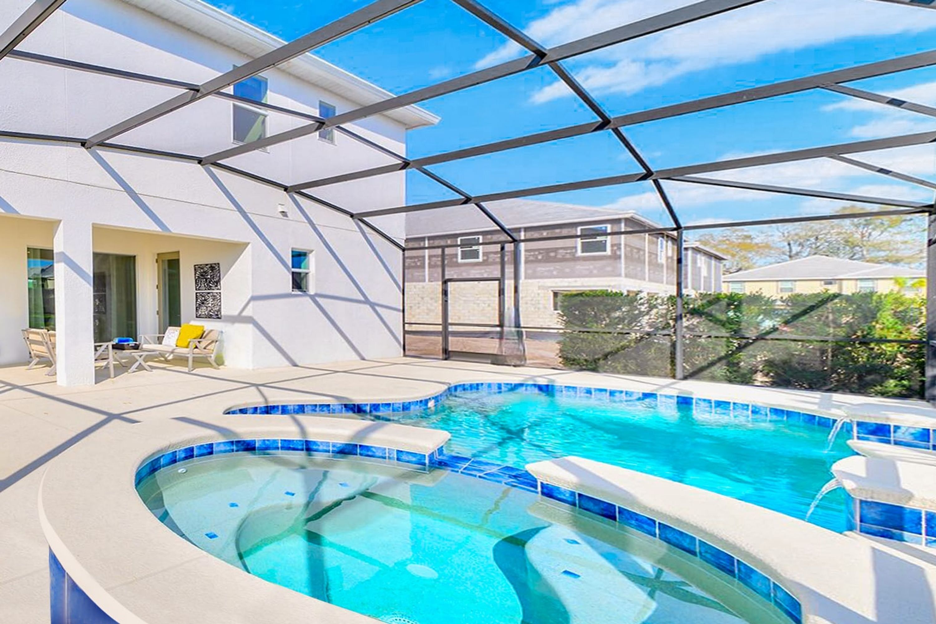 A swimming pool with a glass enclosure, surrounded by a white building with a tiled deck and outdoor seating.