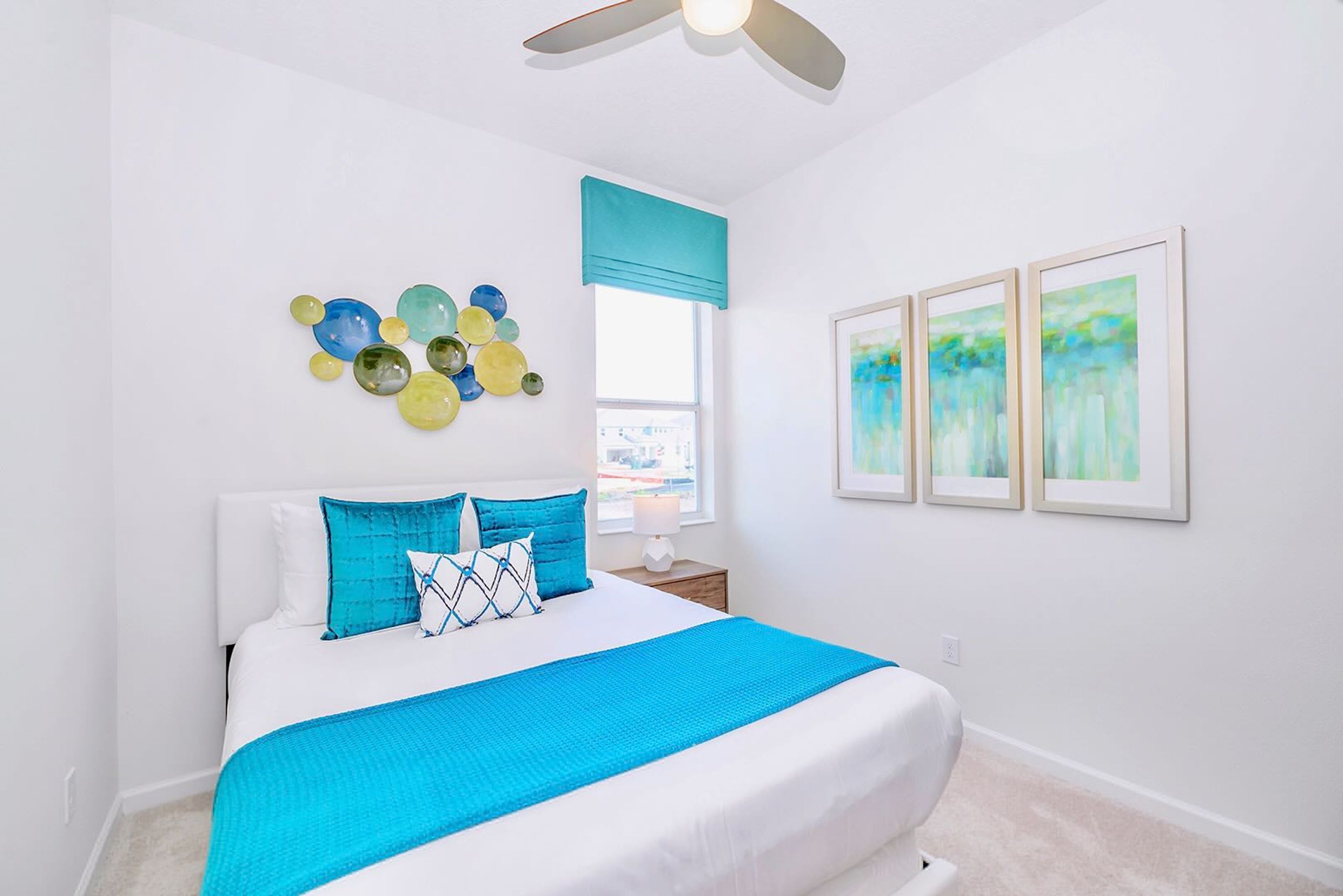 A bedroom with a bed, a ceiling fan, a window with a blue valance, and three framed artworks on the wall.
