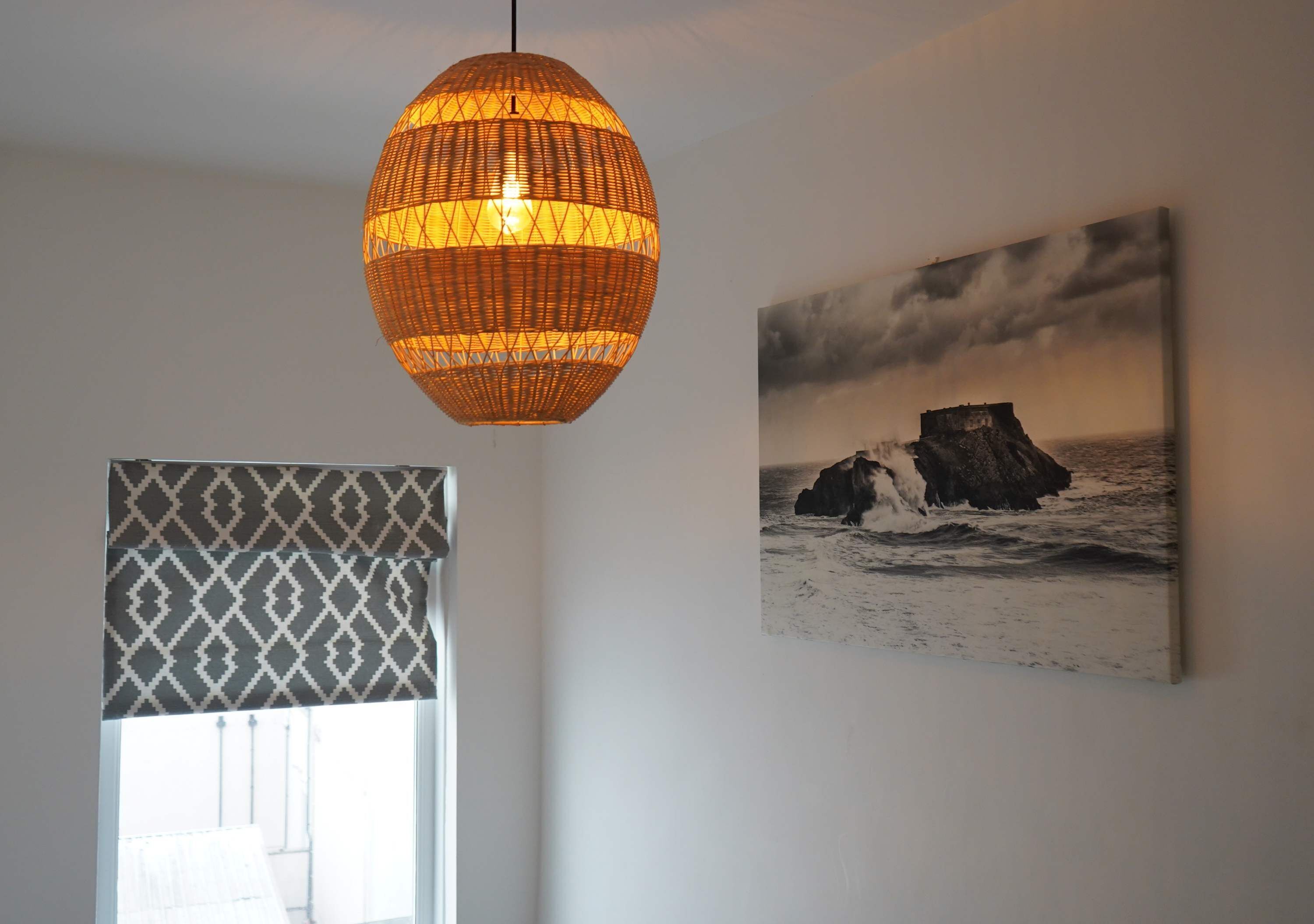 A room with a large woven pendant light, a patterned window valance, and a framed seascape painting.