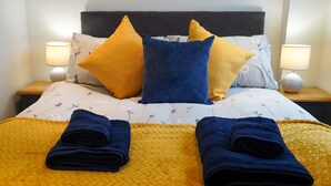 A neatly made bed with a yellow quilt, two blue towels, and a variety of pillows in different colors and patterns. - Coppet Cove 2 Bedroom Apartment - Tenby (Saundersfoot)