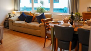 A cozy living room with a sofa, dining table, chairs, and a window with a view of a building. - Coppet Cove 2 Bedroom Apartment - Tenby (Saundersfoot)