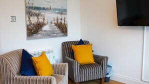 A living room with striped sofas, a painting of a beach scene, and a flat-screen TV mounted on the wall. - Coppet Cove 2 Bedroom Apartment - Tenby (Saundersfoot)