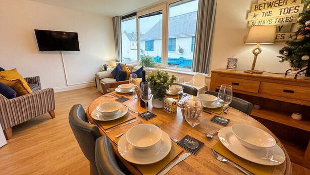 A dining area with a round table set for four, a flat-screen TV mounted on the wall, and a window with a view of a house. - Coppet Cove 2 Bedroom Apartment - Tenby (Saundersfoot)
