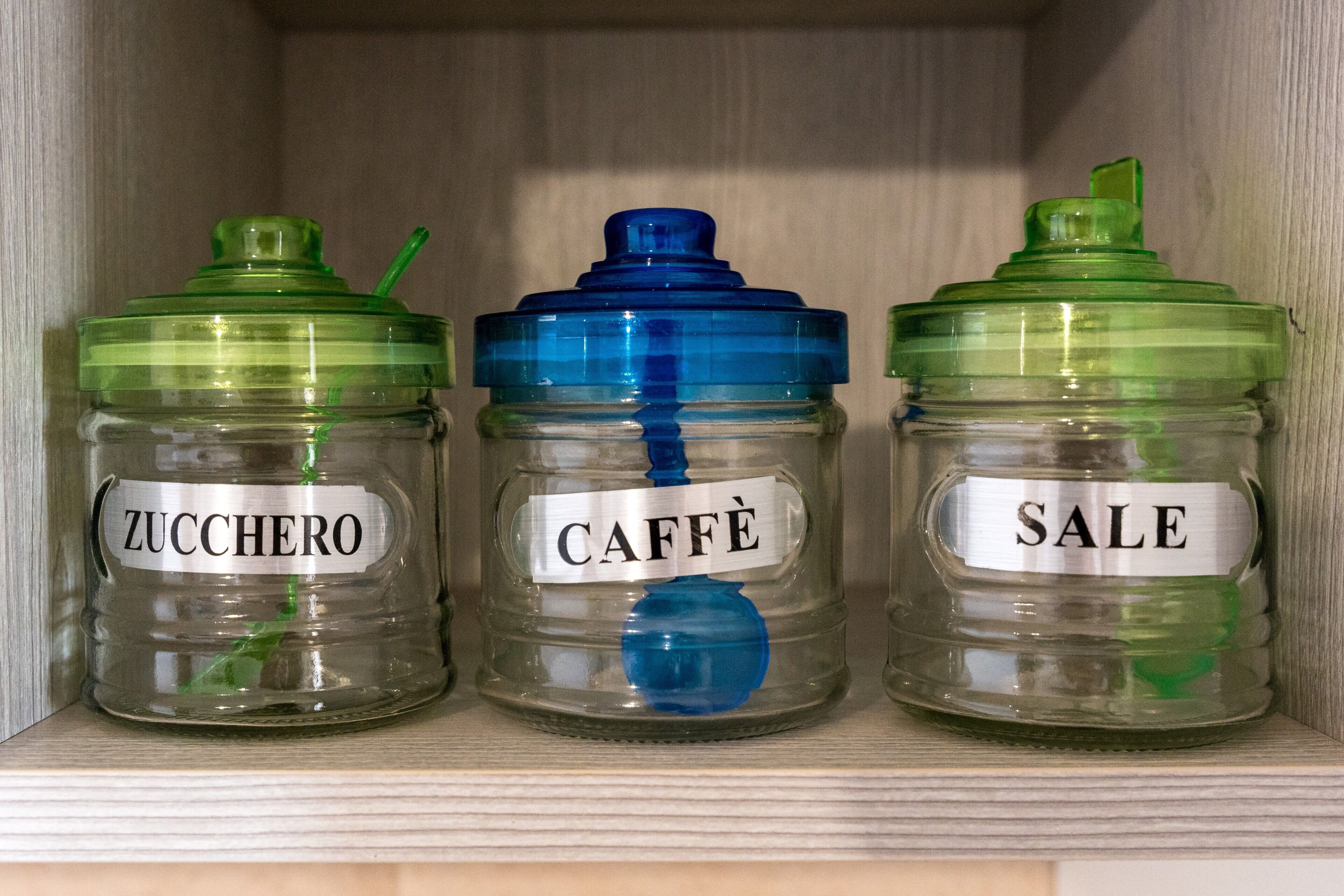 Tres frascos de vidrio con tapas en un estante. Cada frasco tiene una etiqueta con un nombre: 'ZUCCHERO', 'CAFFÈ' y 'SALE'.