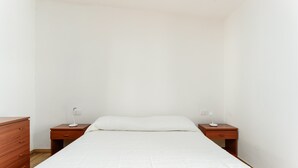 Een eenpersoonsbed met een witte matras en kussens, aan weerszijden voorzien van houten nachtkastjes met bedlampjes.
