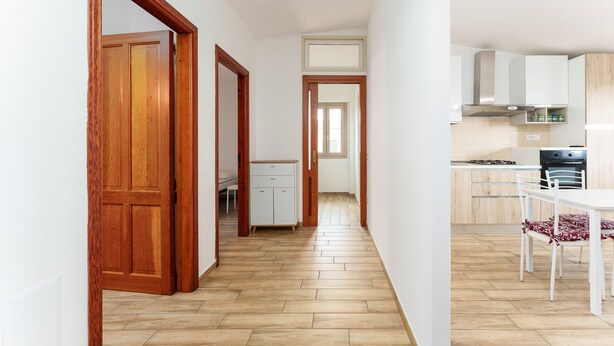 Een smalle gang met houten deuren, een keuken met witte kastjes en een eettafel met stoelen.