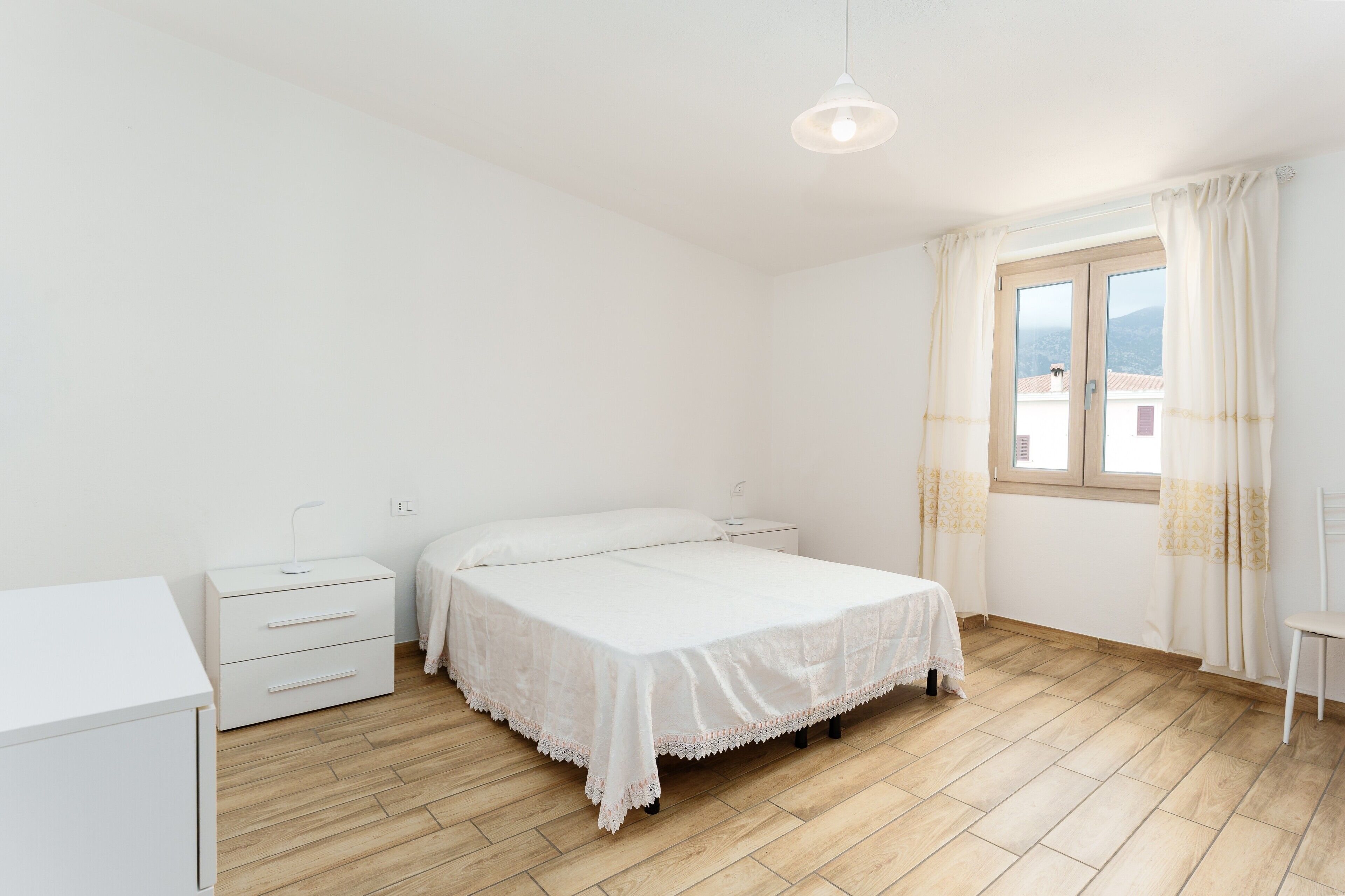 Een slaapkamer met een bed, nachtkastje, stoel, raam met gordijnen en uitzicht.