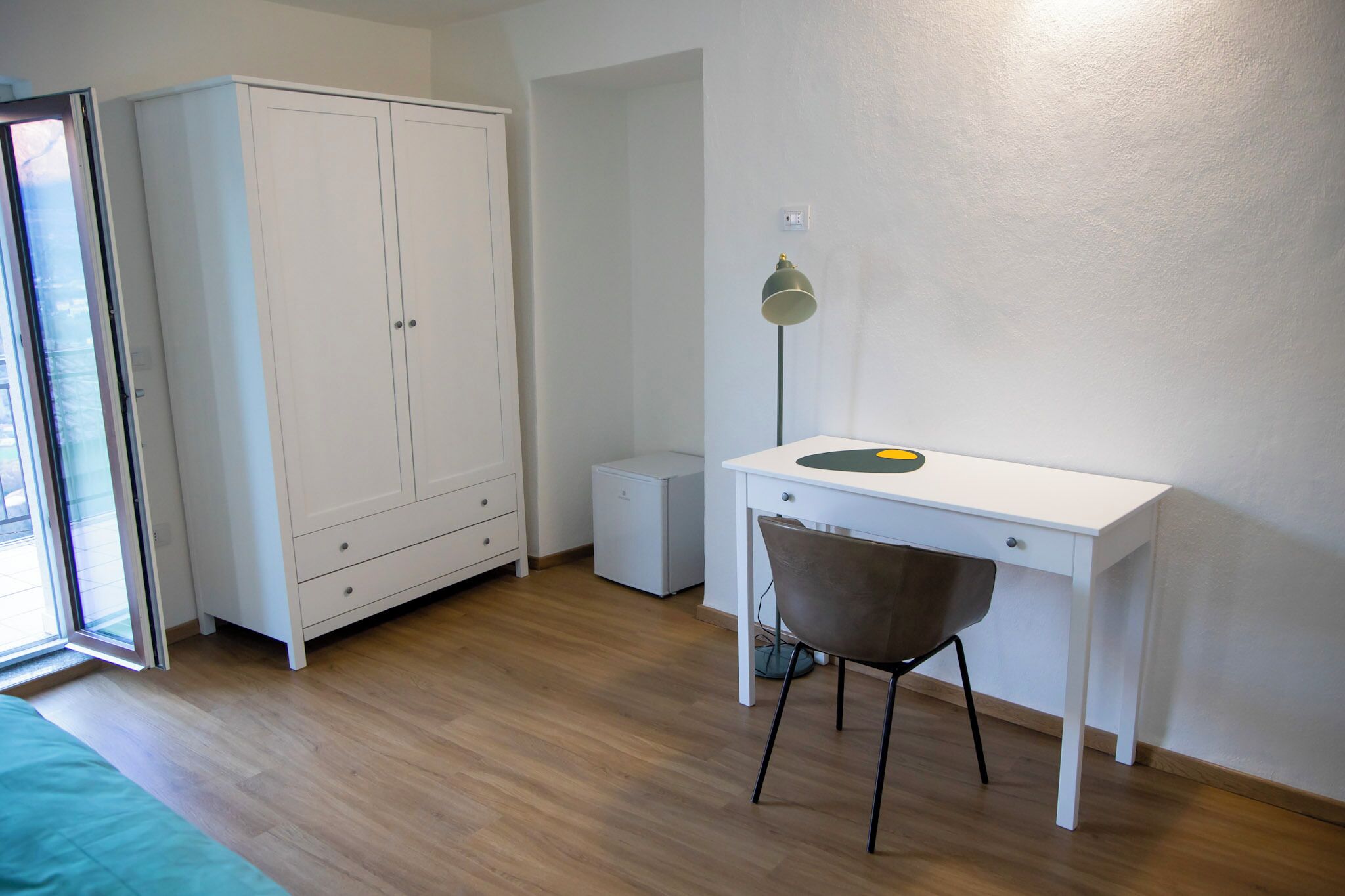A bedroom with a bed, a wardrobe, a desk with a chair, a lamp, and a small bedside table.