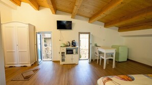 A room with wooden beams on the ceiling, a television mounted on the wall, a kitchenette with a microwave, a small refrigerator, a white desk, and a bed with a colorful headboard. - B&B Anele (Morano Calabro)