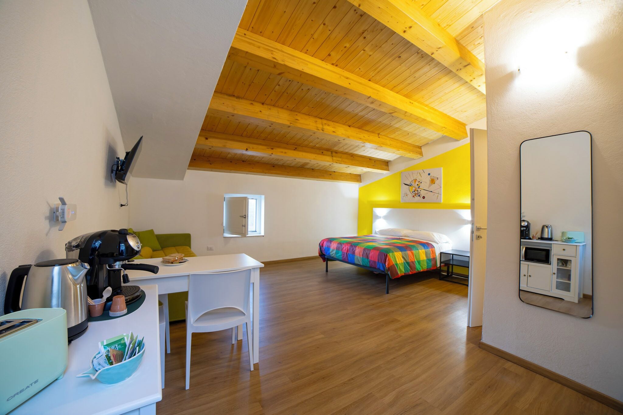 Une pièce avec un plafond en bois, un lit recouvert d’une courtepointe aux couleurs vives, un bureau avec une machine à café et une petite kitchenette.