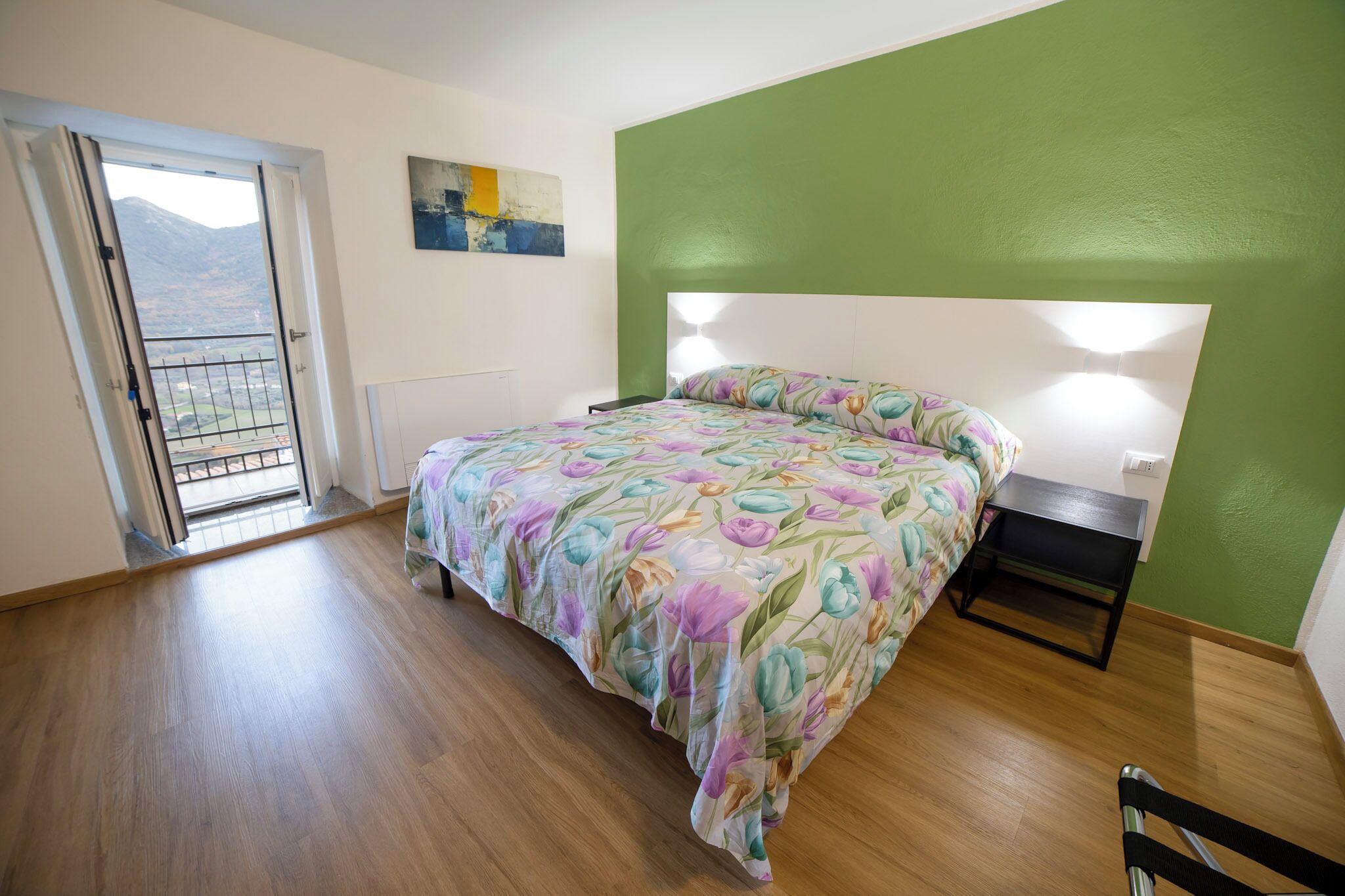 Quarto com cama, mesa de cabeceira, uma cadeira, um quadro e vista através de uma porta.