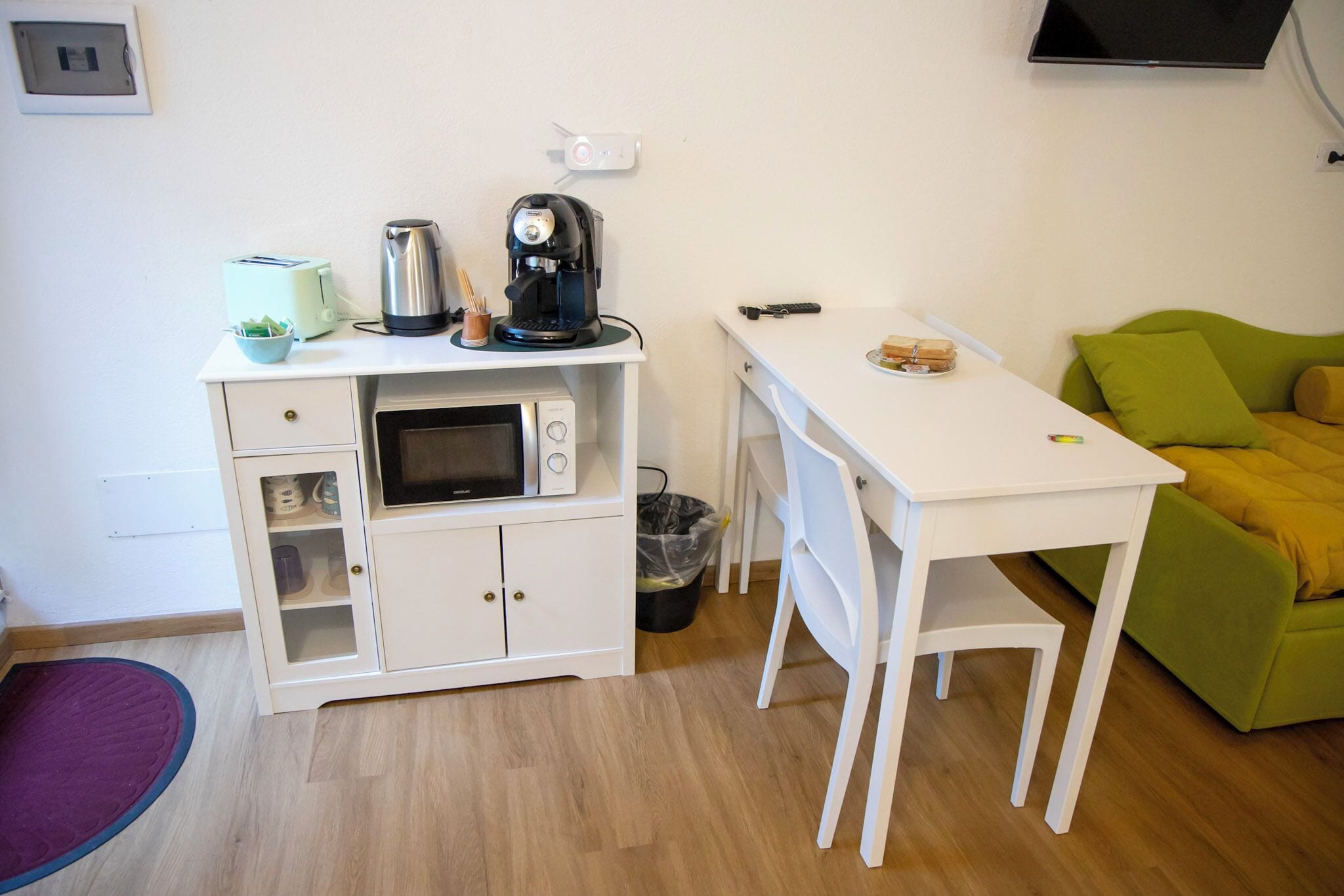 Une petite cuisine équipée d’un four à micro-ondes, d’une machine à café et d’un grille-pain. La pièce comprend également une petite poubelle et un canapé vert.
