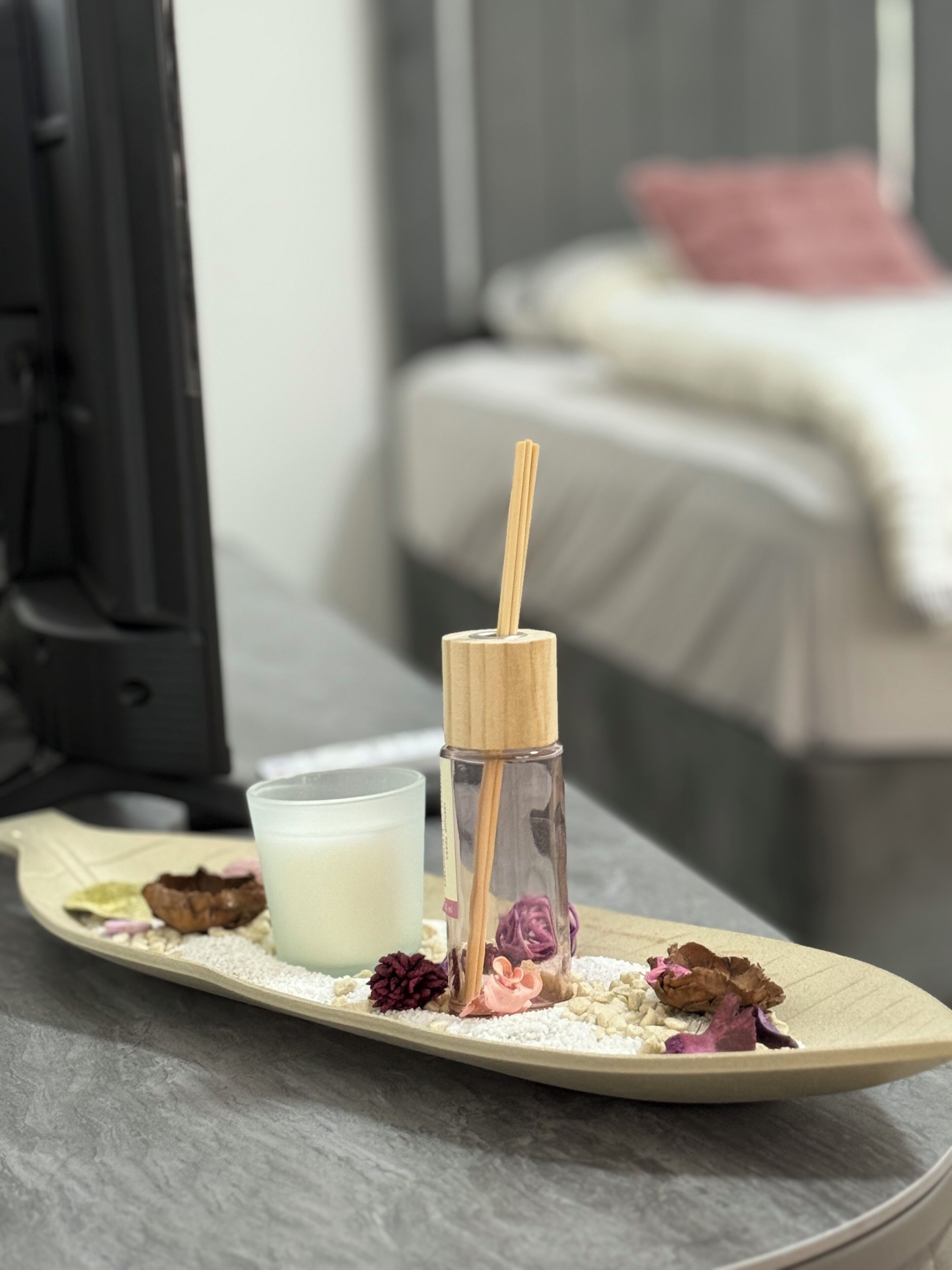 En bakke med et lys, en flaske med låg af træ og nogle tørrede blomster på et bord i et rum med en seng og et fjernsyn i baggrunden.