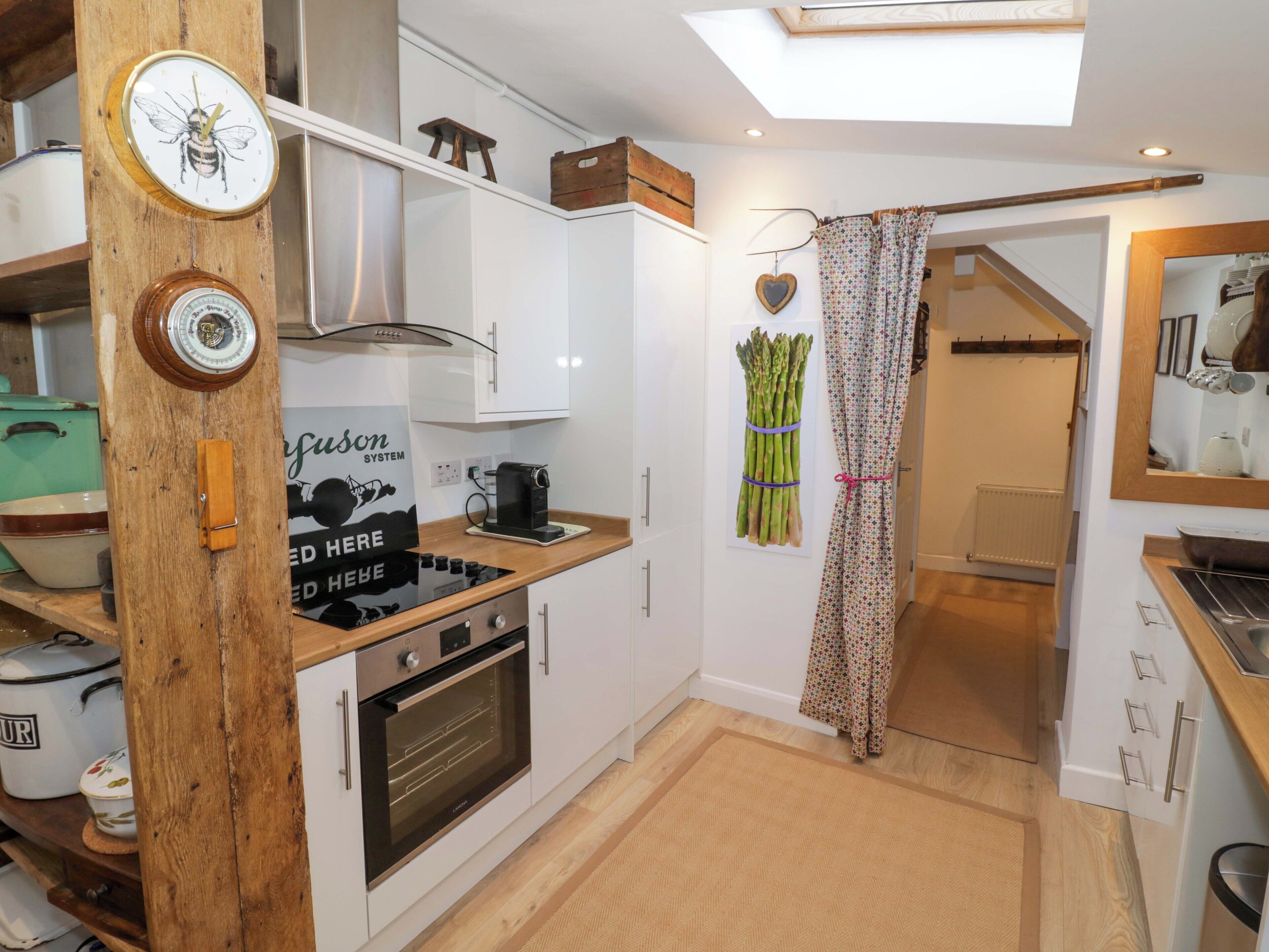 A compact kitchen with a modern stove, oven, and a small coffee machine. There's a wooden beam with a clock and a wall-mounted scale. A staircase leads to another room.