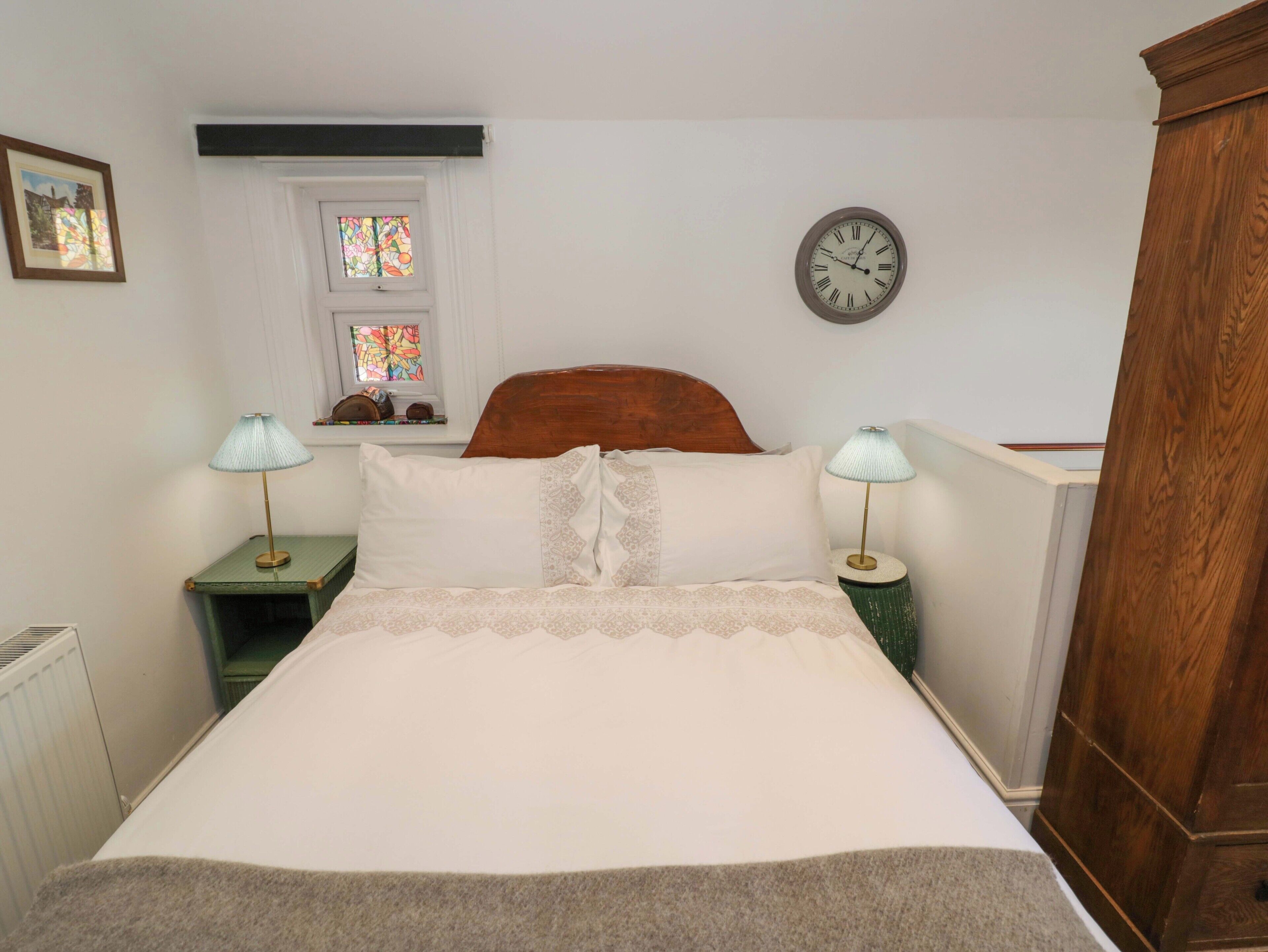 A neatly made bed with a wooden headboard, two bedside tables with lamps, a wall clock, and a window with stained glass.
