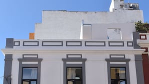 A two-story building with a white facade and gray trim. There is a sign on the ground floor that reads "VENDIDOS DE MAYO DE 1886". - La Isleta Nest Hostel (Las Palmas de Gran Canaria)