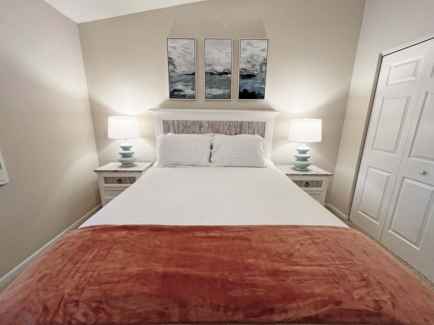 A bedroom with a bed, two bedside tables, a nightstand, a door, and three wall-mounted paintings.