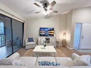 A living room with a ceiling fan, a flat-screen TV, a sofa, a coffee table, and a large window. - Conquistador 8 Navarra Ct (St. Augustine)