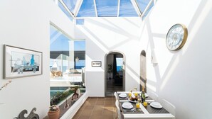 A sunlit dining area with a table set for two, a framed artwork on the wall, and a view of the sea through a glass door. - Puesta Del Sol (Montaña Roja)
