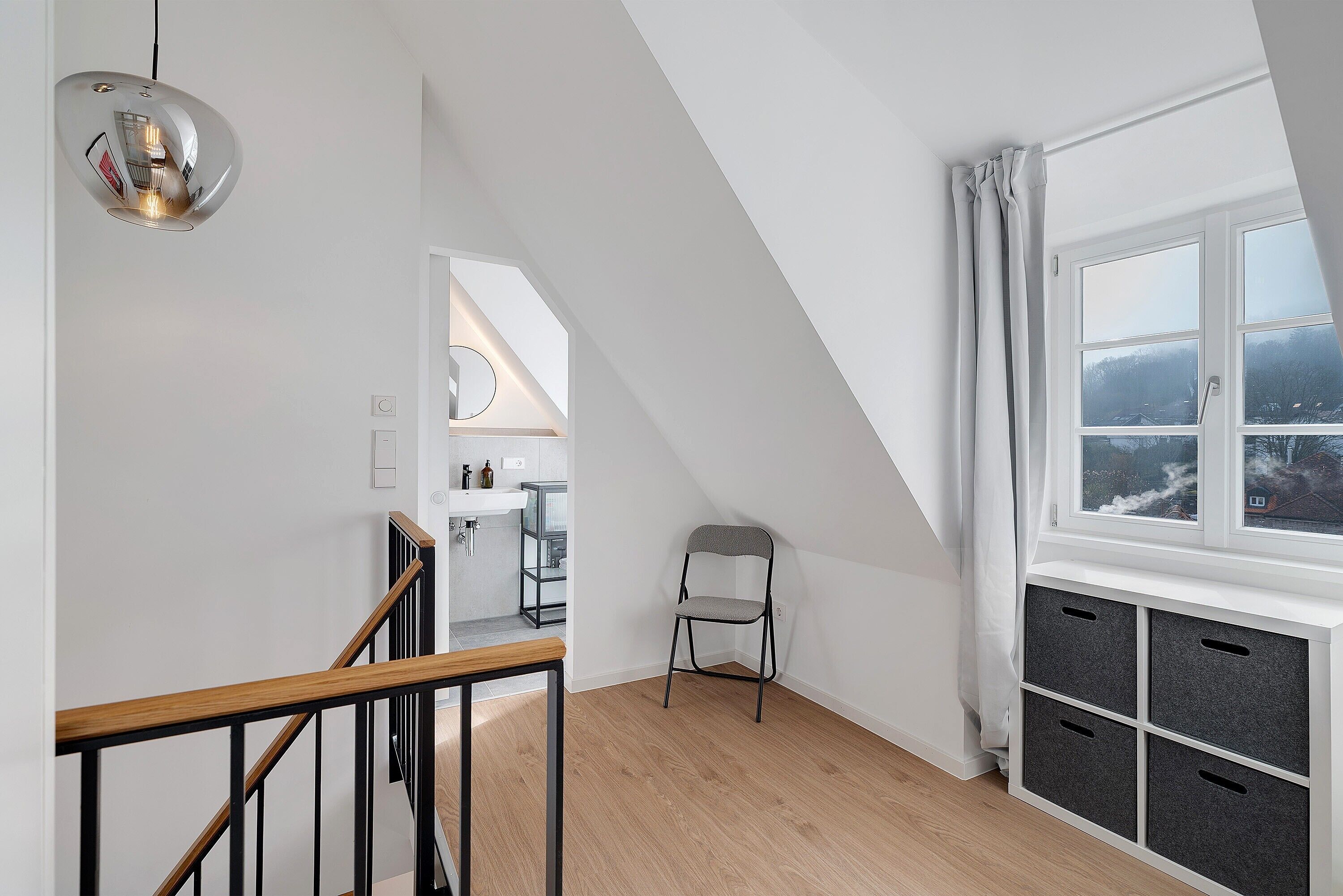 Quarto de loft moderno com teto inclinado, claraboia, escada de madeira e vista para uma paisagem nevada.
