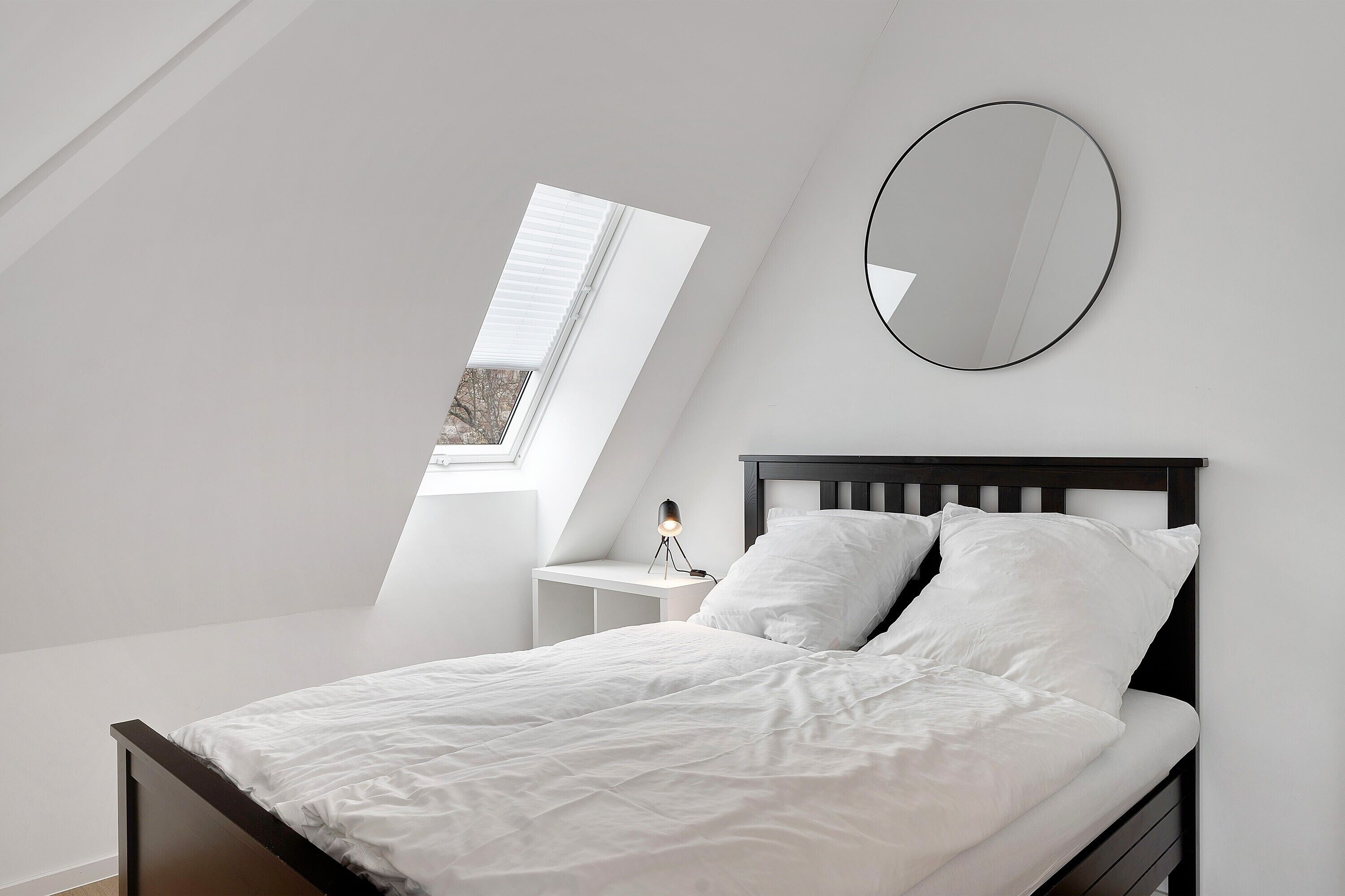 A bedroom with a bed, a bedside table, a lamp, a window, and a wall-mounted mirror.
