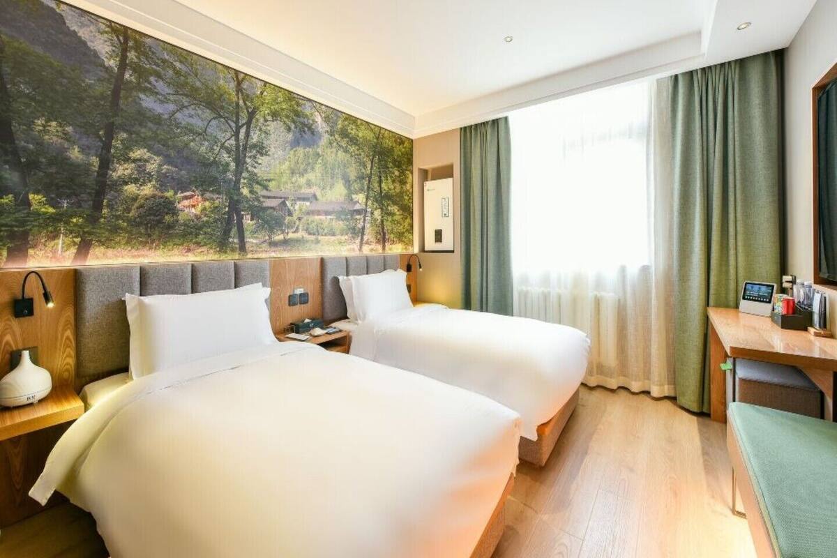 Habitación de hotel con dos camas, un escritorio, una silla, una mesita de noche, una lámpara y un ventanal grande con cortinas.