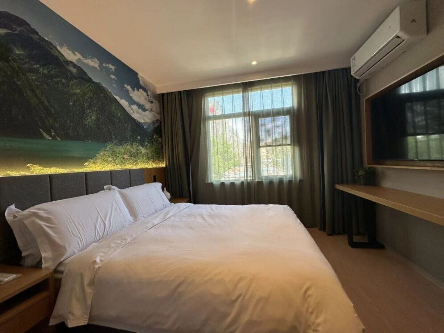 Une chambre d’hôtel avec un lit, une grande fenêtre avec des rideaux, un climatiseur fixé au mur et un tableau représentant un paysage montagneux.